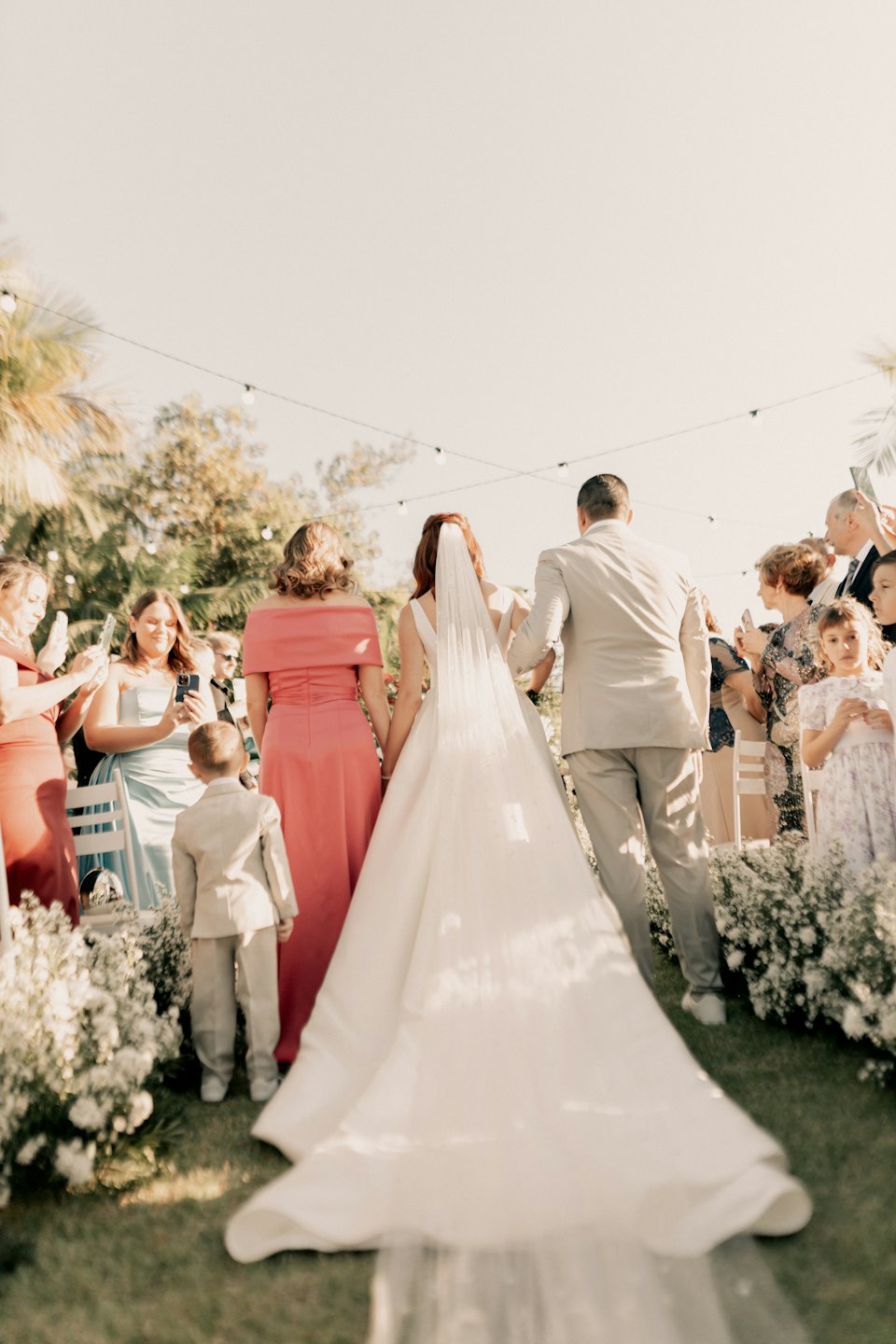 A bride walks down an outdoor aisle with two companions.